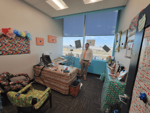 Office decorated with colorful gift wrap covering furniture and walls. A person stands smiling near the desk, with large windows in the background showing a view outside.
