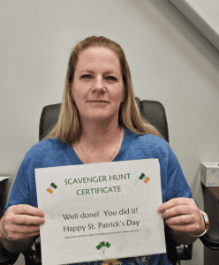 Woman holding a "Scavenger Hunt Certificate" that reads "Well done! You did it! Happy St. Patrick’s Day" and features shamrock decorations.