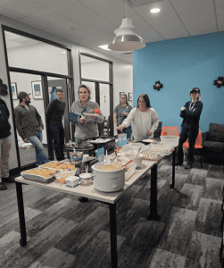People serving food from tables with various dishes in an office setting. Some individuals are standing and conversing in the background.