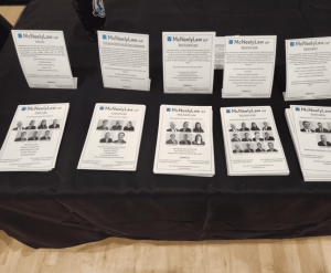 Several stacks of printed flyers and pamphlets are arranged on a black tablecloth, each featuring text and headshots, likely for an informational display or event.