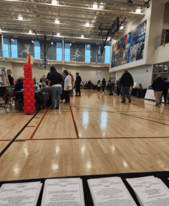 A gymnasium with people gathered at tables for an event, red balloon column on the left, and printed sheets on a table in the foreground.