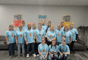 A group of people wearing matching blue T-shirts and decorated masks pose together in an office setting with colorful artwork on the wall behind them.