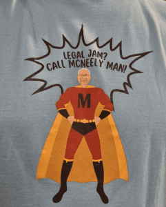 A cartoon superhero with an "M" on his chest and a man's face is printed on a shirt with the text: "Legal Jam? Call McNeely Man!" in a comic-style speech bubble.