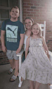 A woman sits on a white rocking chair between a smiling boy in a blue Puma shirt and a girl in a floral dress, with a brick wall and window in the background.