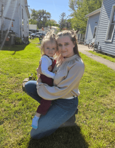 A woman kneels on grass holding a toddler in her arms; houses, bikes, and trees are visible in the background on a sunny day.