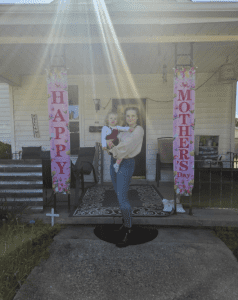 A woman holding a young child stands in front of a house decorated with two pink "Happy Mother's Day" banners and floral designs. Sunlight beams down from above.
