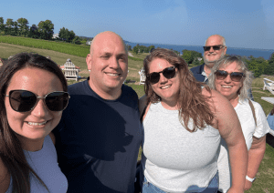 Five adults wearing sunglasses smile outdoors on a sunny day, with green grass, trees, and water visible in the background.