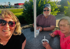Three adults sit around an outdoor table eating ice cream on a sunny day, with greenery and a road visible in the background.