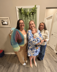 Three women stand indoors, one holding a baby. They are smiling at the camera, with a green and white floral backdrop behind them.