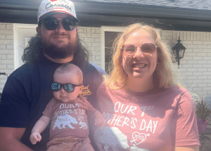 Two adults and a baby wearing sunglasses stand outside a white brick house. The baby and woman wear matching shirts that read "Our 1st Mother's Day 2023.