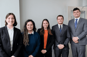 Five people in business attire stand side by side in an office setting, facing the camera and smiling.
