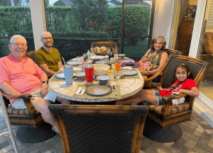 Five people of varying ages sit around a round outdoor table set for a meal, with drinks, plates, and food visible on the table.