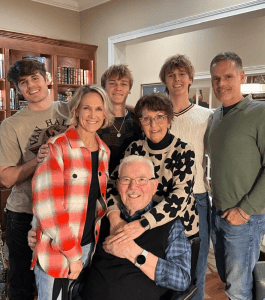 Six people stand and smile around an elderly couple seated in front, posing for a family photo in a warmly lit living room.
