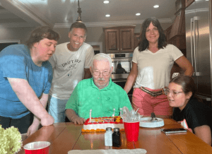An elderly man sits at a table with a birthday cake, surrounded by four people smiling in a kitchen.