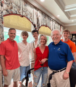 A group of six people—four men, one woman, and one older man—stand together and smile in a well-lit kitchen, with patterned curtains and windows in the background.