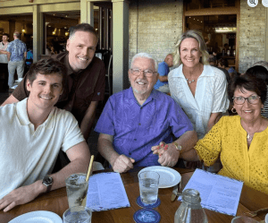 Five people, three men and two women, pose and smile around a restaurant table set with menus, drinks, and water glasses.