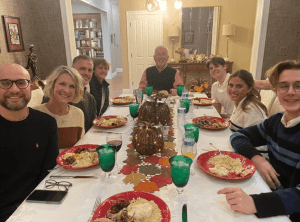 Eight people sit around a dining table set with food, drinks, and fall-themed decorations, smiling at the camera in a warmly lit room.