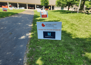 Sign on grass thanks McNeelyLaw as a sponsor for the National Kidney Foundation of Indiana; event setup with people and sponsor signs visible in the background.