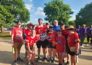 A group of people wearing matching red shirts stand outdoors, holding a sign that says "NKFI Rocks." Trees and other people are visible in the background.