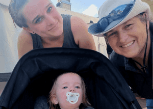 Two adults lean in and smile at the camera, with a baby in a stroller in the foreground sucking on a pacifier.