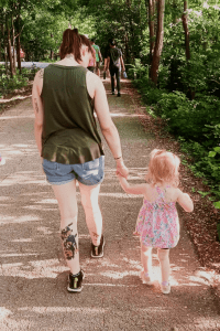 An adult and a young child walk hand-in-hand along a shaded park path, surrounded by greenery on a sunny day.
