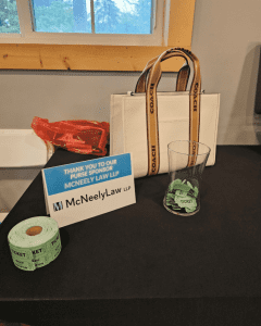 A white Coach handbag sits on a table next to a sponsor sign, a roll of green raffle tickets, and a glass container filled with tickets.