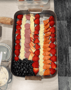 A tray with rows of strawberries, apple slices, salami, cheese, pepperoni, and a pile of blueberries on one side, arranged neatly on parchment paper.