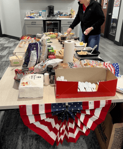 A table in an office kitchen is filled with various foods, snacks, and utensils. A person stands at the far end, and patriotic decorations hang from the table edge.