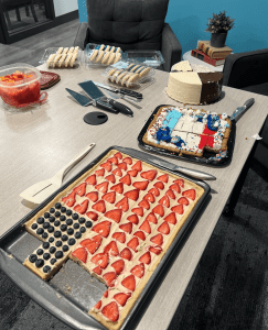 A table with various desserts including an American flag fruit tart, a decorated sheet cake, a layered round cake, cookies, and a bowl of fruit salad.
