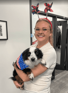 A woman wearing star-and-stripe pinwheel headbands holds a black and white dog dressed in an American flag-themed bandana, standing indoors.