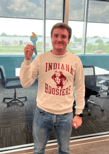 A person wearing an Indiana Hoosiers sweatshirt stands indoors holding a small blue and yellow torch-shaped object.