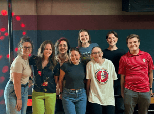 Eight people stand indoors in a casual group, smiling at the camera. The background features a dark wall with red and black horizontal stripes and a few visible red light spots.