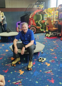 A man in a blue shirt and roller skates sits on a bench in an arcade with colorful carpet; his shoes are on the floor beside him.