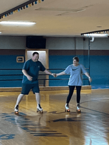 Two people roller skating indoors on a wooden rink, holding hands for balance.