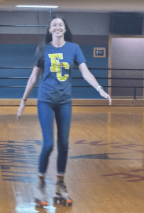 A person wearing a blue "EC" t-shirt roller skates indoors on a wooden rink, smiling with arms outstretched.