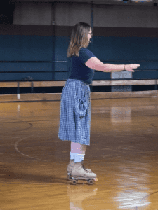 A person wearing a blue shirt, plaid skirt, and roller skates is skating on an indoor rink with wooden flooring.