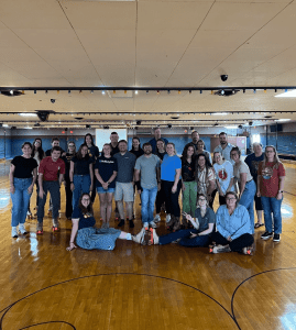 A group of people poses together on a wooden roller skating rink, some standing and some seated on the floor, all wearing roller skates.