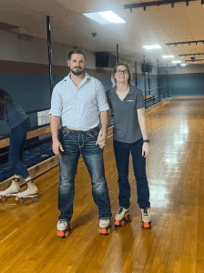 Two people stand side by side on roller skates in an indoor skating rink, holding hands and facing the camera. Another person skates in the background.