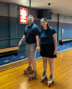 Two people holding hands and roller skating indoors on a wooden rink, both wearing casual clothes and brown skates.