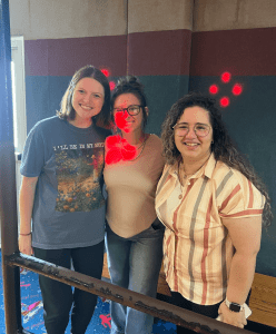 Three women stand together indoors, smiling at the camera. Red digital heart doodles are drawn above and on the woman in the center.