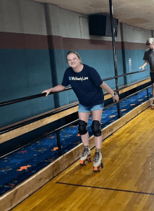 A woman wearing knee pads and a "McNeelyLaw" t-shirt roller skates indoors while holding onto a railing.