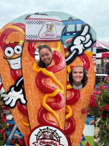 Two people smile through cut-out photo stands shaped like cartoon hot dogs with ketchup and mustard, at an outdoor event or fair.