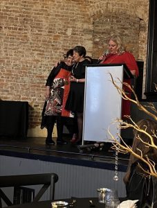 Three women stand on a stage; two are embracing while the third, in red, speaks at a podium. They are in front of a brick wall at an indoor event.