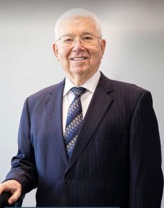 An elderly man wearing glasses and a dark pinstripe suit with a patterned tie, standing and smiling in front of a plain background.