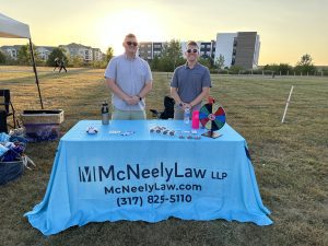 Two people stand behind a table covered with a McNeely Law LLP tablecloth at an outdoor event. Promotional items and a prize wheel are displayed on the table.