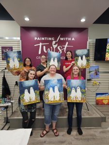 Six women stand on a small stage holding up paintings of ghosts in a field, beneath a sign that reads “Painting With a Twist.”.